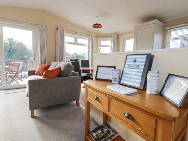 A living room with a grey sofa and orange cushions a wooden table with guest information and framed pictures at Laurel Lodge Bossiney Bay Holiday Park near Tintagel