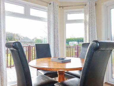 A dining area with a round wooden table and four black leather chairs near windows with curtains at Laurel Lodge Bossiney Bay Holiday Park near Tintagel