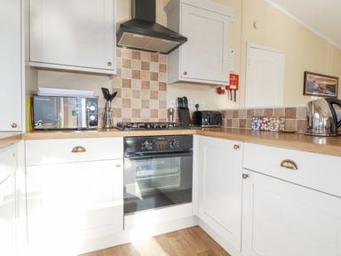 A kitchen with white cabinets a built in oven stove microwave toaster kettle and cooking utensils at Laurel Lodge Bossiney Bay Holiday Park near Tintagel