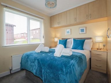 A bedroom with a bed covered in blue bedding and rolled towels on top with bedside tables and lamps at Laurel Lodge Bossiney Bay Holiday Park near Tintagel