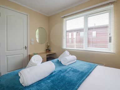 A bedroom with a bed covered by a blue blanket and white rolled towels on top a small wooden desk with a round mirror on the wall and a window with blinds