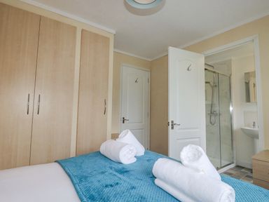 A bedroom with a bed covered in a blue blanket and rolled white towels a wooden wardrobe and an open door leading to a bathroom with a glass shower at Laurel Lodge Bossiney Bay Holiday Park near Tintagel