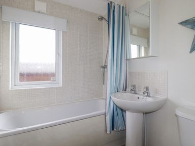 A bathroom with a bathtub a shower with striped curtain a sink with two taps a mirror cabinet and a frosted window at Laurel Lodge Bossiney Bay Holiday Park near Tintagel