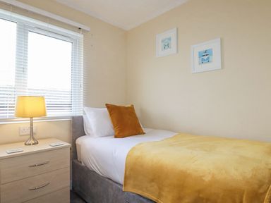 A bedroom with a single bed yellow blanket a pillow a bedside table with a lamp and two framed pictures on the wall at Laurel Lodge Bossiney Bay Holiday Park near Tintagel