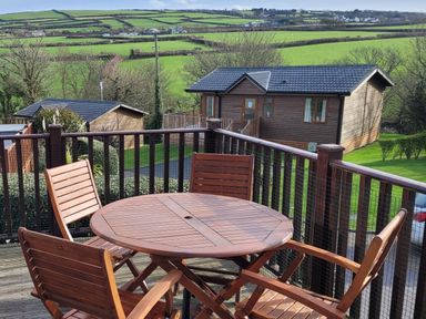A wooden table with four chairs on a balcony overlooking wooden cabins and green fields at Laurel Lodge Bossiney Bay Holiday Park near Tintagel