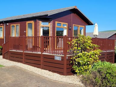 A wooden lodge with a deck and railing surrounded by bushes at Laurel Lodge Bossiney Bay Holiday Park near Tintagel