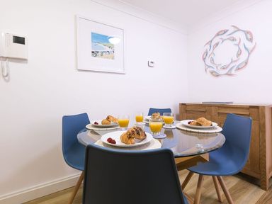 A dining area with a round glass table set with croissants and orange juice and blue chairs at Atlantis in St Ives