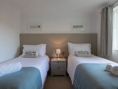 A bedroom with two single beds with a shared headboard two framed pictures and a bedside table with a lamp and flowers at Atlantis in St Ives