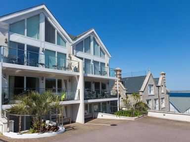 A modern white multi level building with balconies and outdoor chairs next to a stone building by the sea at Atlantis in St Ives