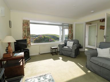 A living room with two gray armchairs a small wooden table a TV on a stand and a large window with floral curtains at 33 Weymouth Park in Hope Cove