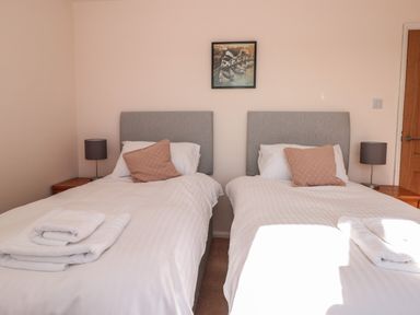 A bedroom with two single beds with grey headboards white bed linens pink cushions bedside tables with lamps and a painting on the wall at 33 Weymouth Park in Hope Cove