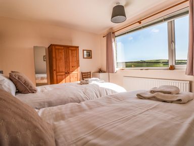 A bedroom with two single beds, wooden wardrobe, chair, large window with curtains overlooking a landscape at 33 Weymouth Park in Hope Cove