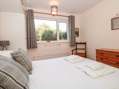 A bedroom with a white bed and folded towels on it a wooden dresser a wooden chair and a window with plaid curtains at 33 Weymouth Park in Hope Cove