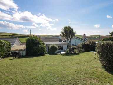 A backyard with grass lawn, bushes, a palm tree, and a small white house with outdoor furniture at 33 Weymouth Park in Hope Cove