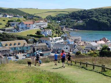 People walking on a grassy path toward a coastal village with buildings and a beach at 33 Weymouth Park in Hope Cove