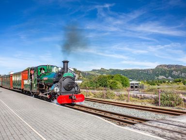 A steam train stopped at a platform with tracks and hills in the background at Harbour View in Porthmadog