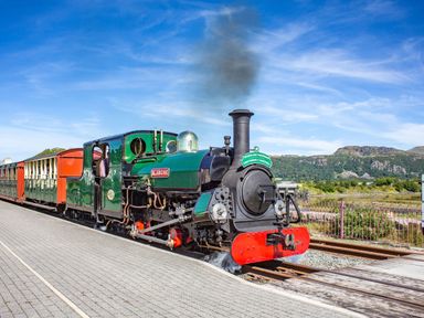 A steam locomotive named Blanche at a railway station with carriages and hills in the background at Harbour View in Porthmadog