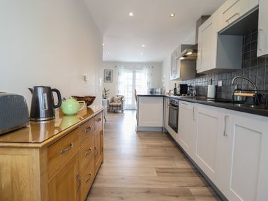 A kitchen with white cabinets a wooden sideboard with a toaster kettle and teapot and a sitting area with armchair at Harbour View in Porthmadog