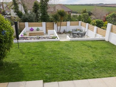 A backyard with a grass lawn a patio seating area with a table and chairs and a wooden fenced garden at Estuary View in Newquay