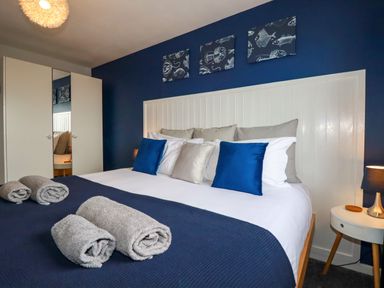 A bedroom with a white bed headboard blue pillows folded towels and a bedside table with a lamp at Estuary View in Newquay