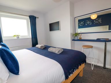 A bedroom with a bed towels on top a window with curtains a wall-mounted television a mirror above a blue shelf with a plant and a stool at Estuary View in Newquay
