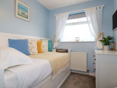 A bedroom with a bed with cushions and a blanket a window with curtains a white dresser and a plant at Estuary View in Newquay