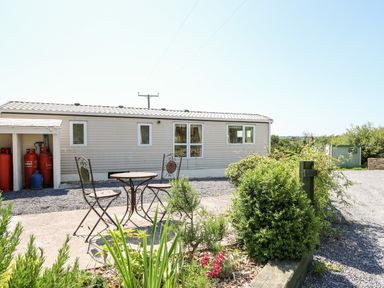 A metal table with two chairs on a patio next to a beige mobile home with propane tanks and greenery at The Lodge in Pontyates