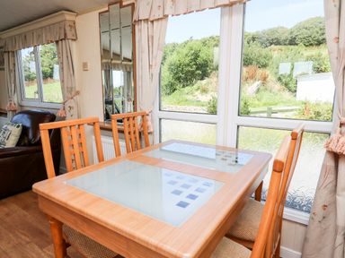 A dining area with wooden table and chairs near large windows and curtains with a view outside at The Lodge in Pontyates
