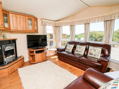 A living room with leather sofas, cushions, a television on a wooden stand, a fireplace, large windows with curtains, and a white rug on the wooden floor at The Lodge in Pontyates