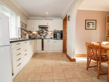 A kitchen with white cabinets a tiled floor and a wooden dining table with chairs at Hazel Cottage in Tintagel