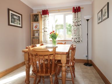 A dining room with a wooden table and chairs a vase of flowers on the table a window with patterned curtains a corner shelf and framed pictures on the walls at Hazel Cottage in Tintagel