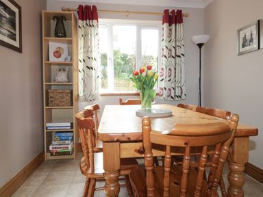 A dining room with a wooden table and chairs a vase of flowers on the table shelves with books and decorations and a tall floor lamp at Hazel Cottage in Tintagel