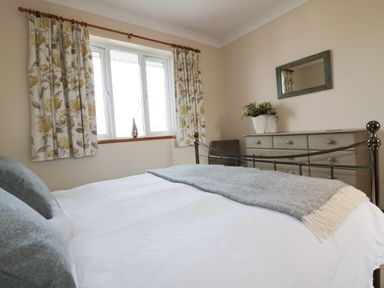 A bedroom with a bed white bed linens a gray blanket and pillows a window with floral curtains a dresser a mirror and a vase with flowers at Hazel Cottage in Tintagel