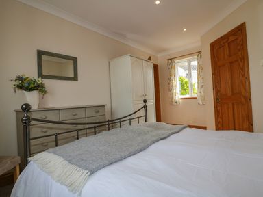 A bedroom with a metal bed frame, dresser with a flower vase, wardrobe, window with curtains, and wooden doors at Hazel Cottage in Tintagel