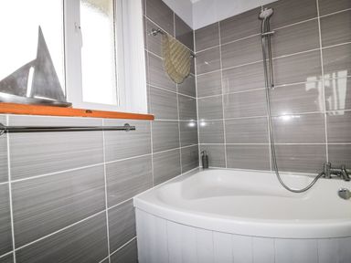 A bathroom with a white bathtub shower mixer and showerhead grey tiled walls and a window with a metal sailboat decoration on the sill at Hazel Cottage in Tintagel