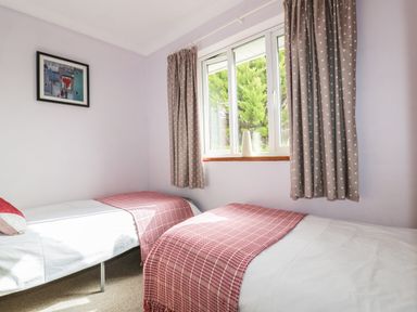 A bedroom with two single beds with red throws near a window with curtains and a framed picture on the wall at Hazel Cottage in Tintagel