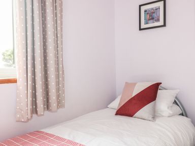 A single bed with pillows and a red and pink cushion beside a window with polka dot curtains and a framed picture on the wall at Hazel Cottage in Tintagel