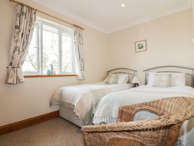 A bedroom with two single beds a wicker chair a window with curtains and a lamp at Hazel Cottage in Tintagel