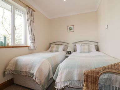 A bedroom with two single beds a bedside table a lamp a window with curtains and a wicker chair at Hazel Cottage in Tintagel
