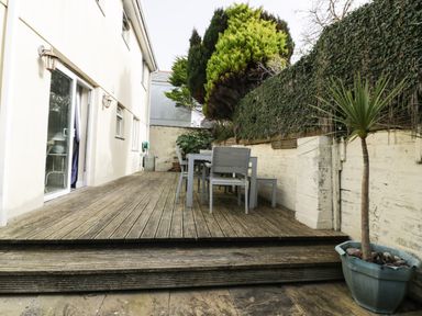 An outdoor wooden deck with a dining table and chairs next to a building with sliding doors and a potted plant on a patio at Hazel Cottage in Tintagel