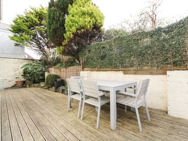 An outdoor wooden deck with a table and four chairs surrounded by plants and a hedge at Hazel Cottage in Tintagel
