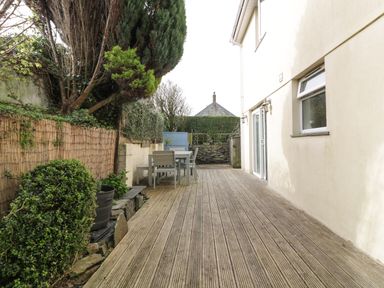 An outdoor wooden deck with patio furniture and plants next to a cream house wall at Hazel Cottage in Tintagel