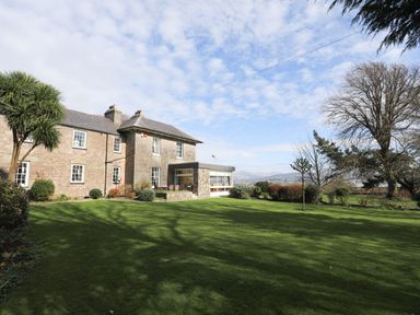 A large stone building with multiple windows surrounded by a lawn and trees at Tal Gwynedd in Dwyran near Newborough