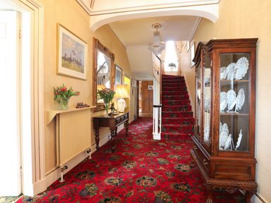 A hallway with a red patterned carpet wooden console table with lamp and flowers mirror and cabinet with plates at Tal Gwynedd in Dwyran near Newborough