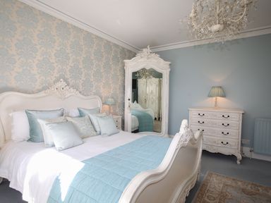 A bedroom with a white carved bed frame a blue quilt and pillows a white dresser with a lamp a large white mirror and patterned wallpaper at Tal Gwynedd in Dwyran near Newborough