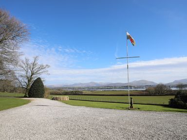 A gravel driveway leading to a grassy area with a flagpole flying a flag and mountains in the background at Tal Gwynedd in Dwyran near Newborough