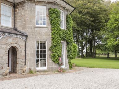 The exterior of a stone house with ivy on the walls two dog statues near an arched doorway and a lawn with trees at Tal Gwynedd in Dwyran near Newborough