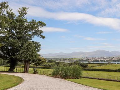 A gravel driveway curving past trees and bushes with fields water and mountains in the background at Tal Gwynedd in Dwyran near Newborough
