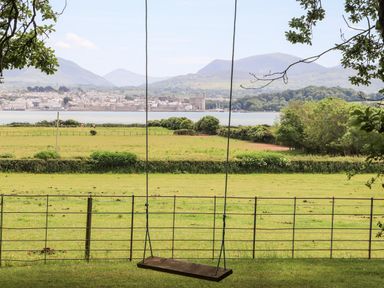A swing hanging from a tree overlooking fields a body of water and mountains at Tal Gwynedd in Dwyran near Newborough