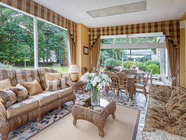 A sunroom with wicker furniture including sofas and tables and large windows showing a garden outside at Tal Gwynedd in Dwyran near Newborough